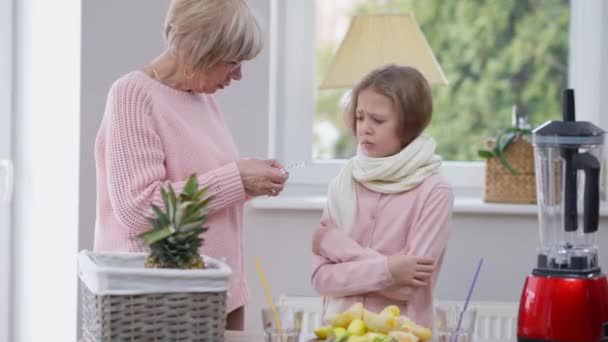 Äldre kvinna med termometer till ledsen flicka vid köksbänk.